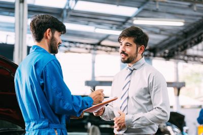 Caucasian automotive maintenance mechanic young man explain car condition to male customer in garage at auto repair shop, technician writing document on vehicle checklist, after service concept